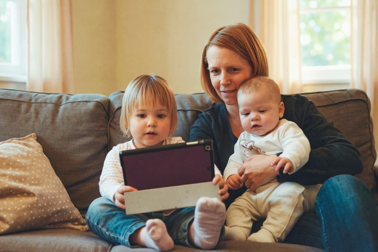 A mother at home on the couch with her newborn and toddler, using a tablet in warm light